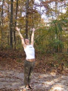 Woman in white shirts and brown pants stands in a forest with her arms stretched up to the sky