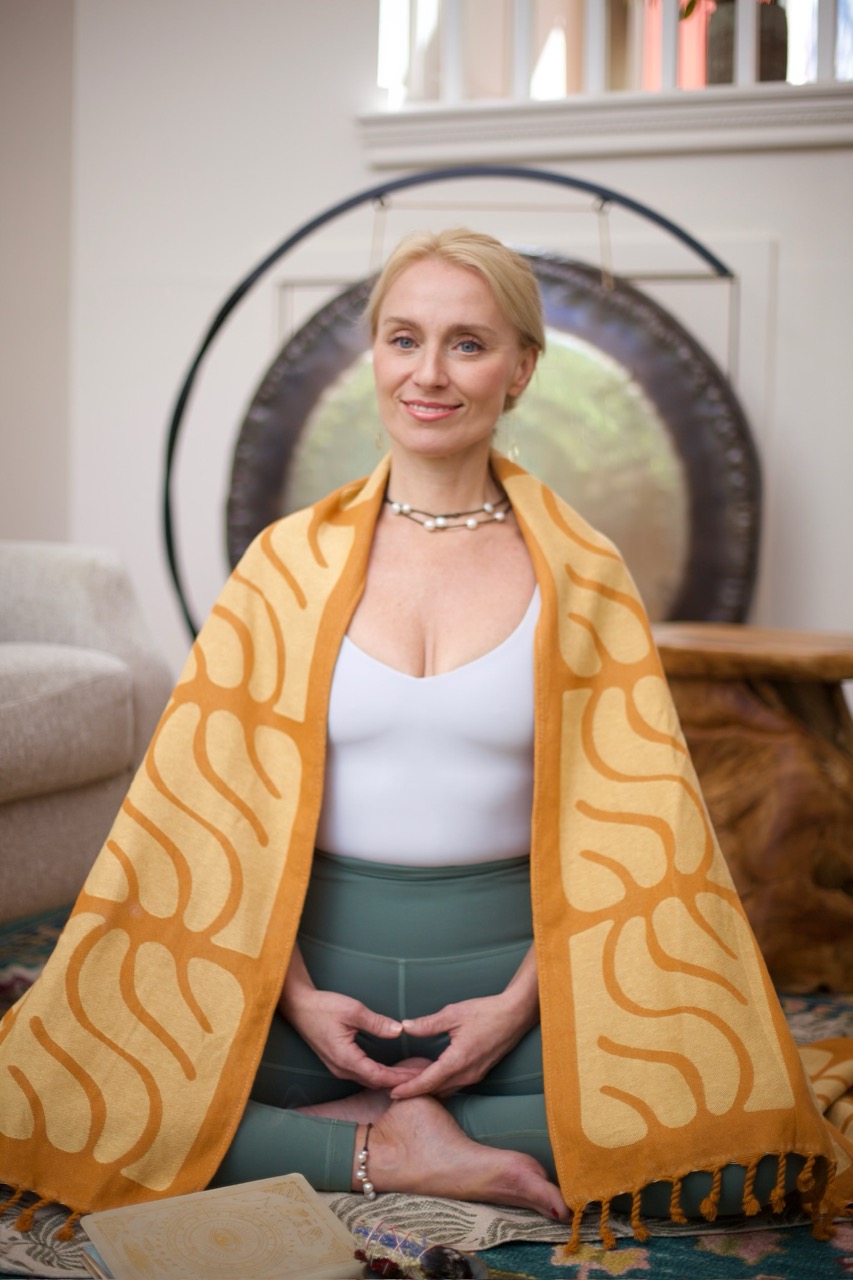 a woman with blond hair and a yellow blanked draped over her back and shoulders is sitting in a cross-legged pose on the floor