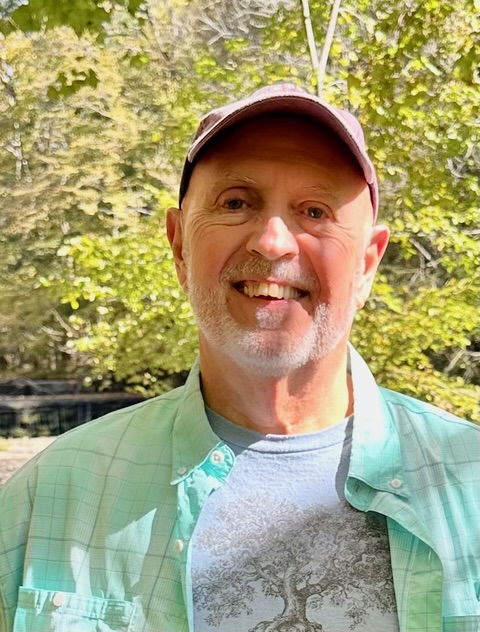 a gray bearded man in a green shirt and hat, smiling, looking at camera. 